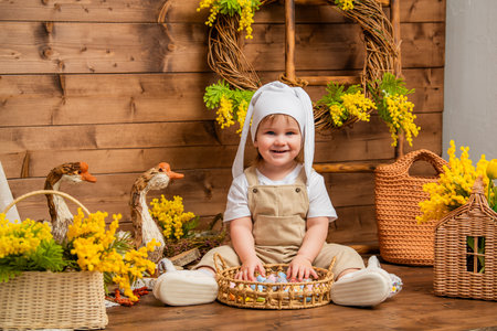 Smiling toddler in bunny hat sitting with basket of easter eggs in rural spring settingの写真素材