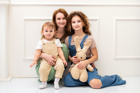 A loving mother sits with her two daughters in a bright, minimalist interior. Their warm expressions and cozy outfits create a touching and tender family momentの写真素材