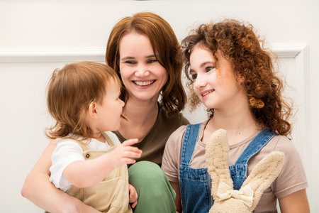 A loving mother sits with her two daughters in a bright, minimalist interior. Their warm expressions and cozy outfits create a touching and tender family momentの写真素材