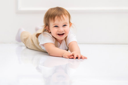 joyful toddler with bright blue eyes and light brown hair lies on a reflective white floor, smiling cheerfully. The child's laughter and natural happiness create a heartwarming and playful sceneの写真素材
