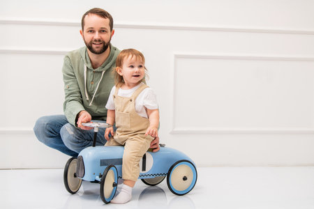 Loving father giving ride to little child in beige overalls and white shirt on blue toy car. Clean minimalist background of white interior.の写真素材