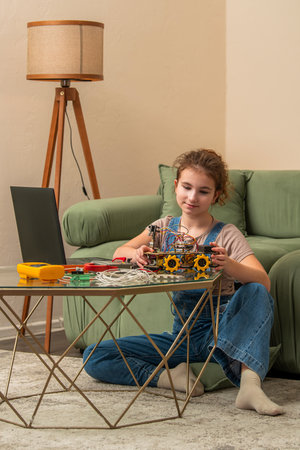 Young girl focused on building a robot model at home, sitting on the floor with a laptop by her side. Tools and wires are scattered on the table, capturing a moment of concentration and learningの写真素材
