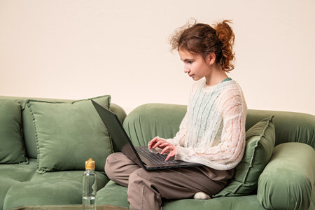 Girl sitting cross-legged on a green couch, focused on working or studying on her laptop. She appears concentrated and engaged with her task, set in a cozy home environmentの写真素材