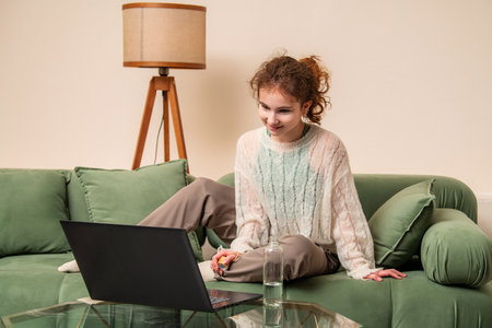 Young girl attentively watches a laptop while sitting on a green couch, holding a bottle of water. She appears relaxed and focused in a cozy home environmentの写真素材