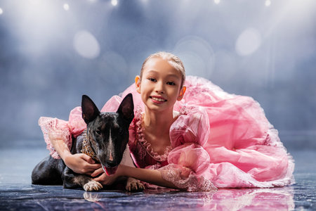 young girl in an elegant pink dress happily hugs a bull terrier on a reflective stage. Both look happy, showing friendshipの写真素材
