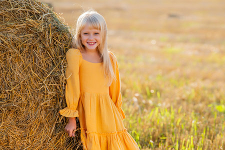 Young girl in a yellow dress leaning against a hay bale on a sunny day. She smiles warmly, capturing the essence of a peaceful rural setting. Ideal for themes of childhood and natureの写真素材
