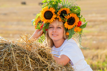 in a bright crown of sunflowers and a yellow dress leaning against a haystack in a sunlit field. Harvest festival.の写真素材