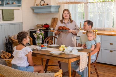 Family gathered around a kitchen table enjoying breakfast. A parent serves fresh pastries to a child while others are seated, creating a warm and cozy family atmosphereの写真素材
