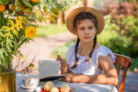 Young girl in a summer dress with braids and hat is seated at a table reading a book outdoors. The scene captures a peaceful moment under the shade of a tree and vivid surroundings.の写真素材