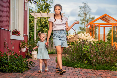 A cheerful moment where an older sister holds a basket and a younger sibling carries a watering can, walking hand in hand along a garden path. Both display joy and companionshipの写真素材