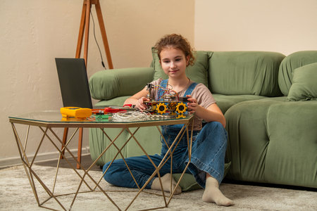 Young girl focused on building a robot model at home, sitting on the floor with a laptop by her side. Tools and wires are scattered on the table, capturing a moment of concentration and learningの写真素材