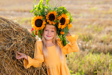 A young girl in a bright crown of sunflowers and a yellow dress leaning against a haystack in a sunlit field. Harvest festival.の写真素材