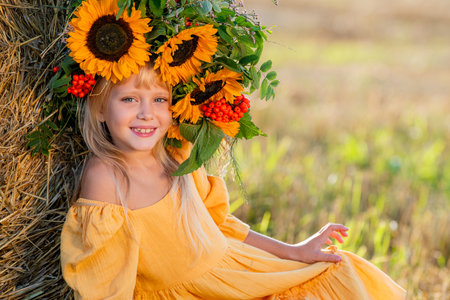 Smiling girl in a yellow dress sits by a haystack in a sunny field, holding a bouquet of sunflowers, radiating joy and warmth in a rural setting surrounded by hayの写真素材