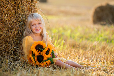 Smiling girl in a yellow dress sits by a haystack in a sunny field, holding a bouquet of sunflowers, radiating joy and warmth in a rural setting surrounded by hayの写真素材