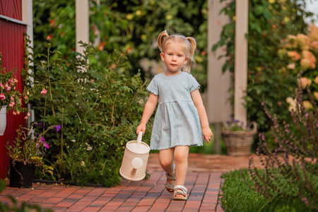 little girl in a blue dress walks along a garden path holding a watering can. Greenery and flowers surround her, creating a warm summer atmosphereの写真素材