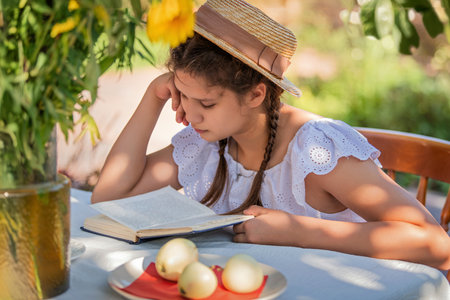 Young girl in a summer dress with braids and hat is seated at a table reading a book outdoors. The scene captures a peaceful moment under the shade of a tree and vivid surroundings.の写真素材