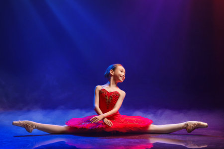Little Ballerina gracefully dances in pointe shoes on stage in bright red tutu against dramatic spotlight backgroundの写真素材