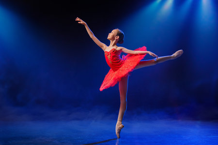 Little Ballerina gracefully dances in pointe shoes on stage in bright red tutu against dramatic spotlight backgroundの写真素材