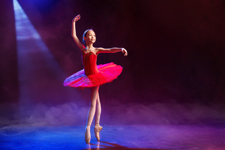 Little Ballerina gracefully dances in pointe shoes on stage in bright red tutu against dramatic spotlight backgroundの写真素材