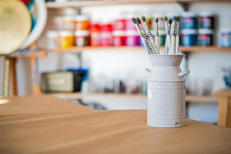 Paintbrushes neatly arranged in a white can on a studio table, surrounded by colorful paint jars in the background, creating an inspiring artistic workspace atmosphereの写真素材