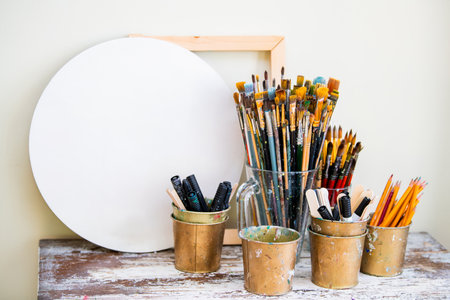 Art supplies including a large round canvas, various brushes in a clear container, and metal cups filled with pencils and markers arranged on a rustic table, ready for creative useの写真素材