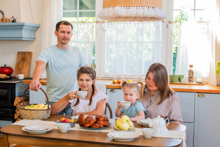 Family gathered around a kitchen table enjoying breakfast. A parent serves fresh pastries to a child while others are seated, creating a warm and cozy family atmosphereの写真素材