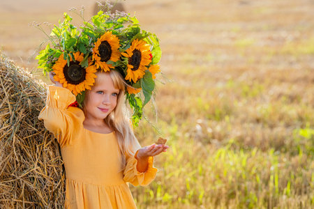 A young girl in a bright crown of sunflowers and a yellow dress leaning against a haystack in a sunlit field. Harvest festival.の写真素材