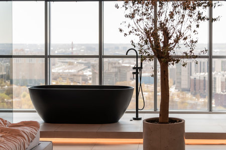 Spacious bathroom featuring a sleek black bathtub next to a large window with a panoramic city view. Includes a potted tree and modern faucet, creating a serene ambiance.の写真素材