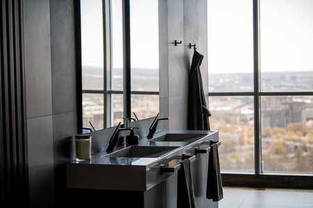 Contemporary bathroom interior featuring double sinks, sleek faucets, and dark towels, against a backdrop of large windows showing an expansive city skyline view.の写真素材