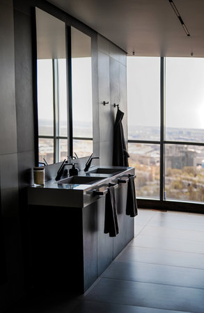 Sleek modern bathroom featuring a double vanity with black fixtures. Expansive windows offer a panoramic city view, enhancing the luxurious and minimalist design aesthetic.の写真素材