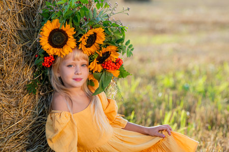 Smiling girl in a yellow dress sits by a haystack in a sunny field, holding a bouquet of sunflowers, radiating joy and warmth in a rural setting surrounded by hayの写真素材