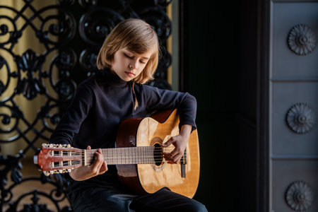 A young boy sits on a stool and plays the guitar. He's wearing a black sweater and he's focused on his music.の写真素材