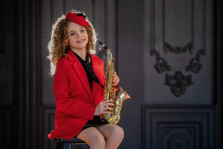 Young girl in fashionable attire sits confidently playing a saxophone in a studio. Dressed in a red beret and jacket with a lively expression, she exudes joy and talentの写真素材