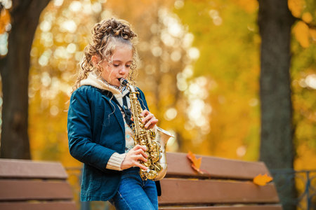 Little girl playing saxophone in autumn park on sunny day. Happy child practice music instrument outdoors for music lesson.の写真素材