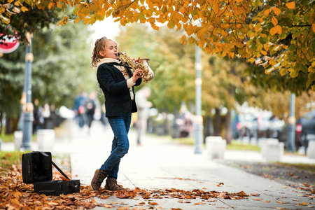 Young girl playing saxophone on street side with fallen autumn leaves around. Music performance concept in city park during Fall season.の写真素材