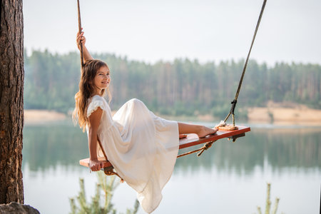 girl in the forest swinging on a swing. Rope swing on a forest lake. Barefoot girl in a white dress with long hair.の写真素材