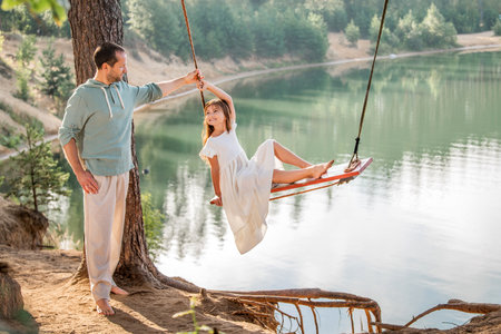 father swings his daughter on a swing on the shore of a forest lake.の写真素材