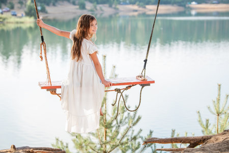 girl in the forest swinging on a swing. Rope swing on a forest lake. Barefoot girl in a white dress with long hair.の写真素材