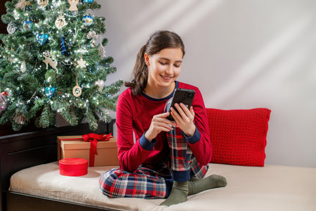 Smiling woman in red pajamas using mobile phone near Christmas tree and gifts. Happy holiday season morning concept.の写真素材