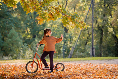 Little girl riding kick scooter through yellow leaves in autumn park. Child playing outdoor during Fall season, childhood concept.の写真素材