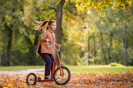 A young caucasian woman riding a scooter on a path covered with autumn leaves in a park during sunny day. Autumn activity concept.の写真素材
