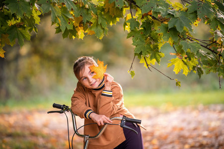 A young girl on a bicycle covering her eye with a vibrant orange autumn leaf in an outdoor park setting. Childhood fun concept for fall season.の写真素材