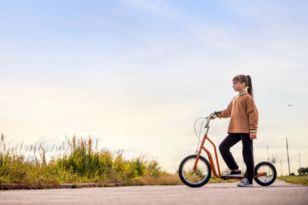 A young girl stands with her orange kick scooter on an empty road next to a grassy area, looking thoughtful.の写真素材