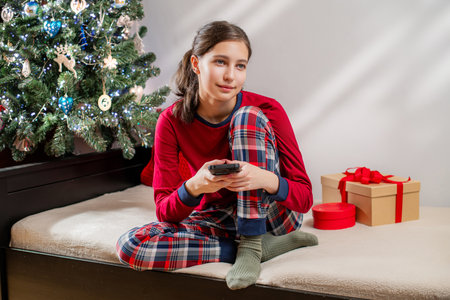 Smiling woman in red pajamas using mobile phone near Christmas tree and gifts. Happy holiday season morning concept.の写真素材
