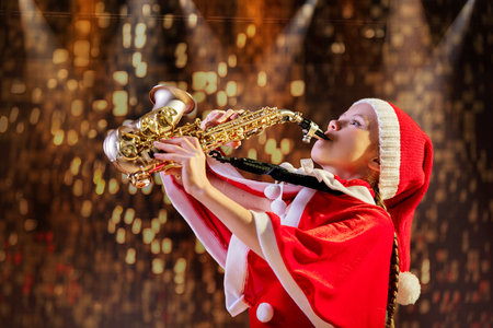 Girl child in red Christmas Santa hat playing saxophone in decorated hall. Christmas music concept.の写真素材