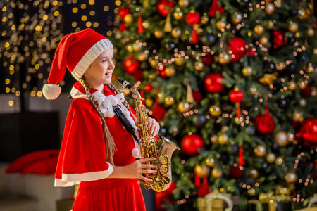 Girl child in red Christmas Santa hat playing saxophone near Christmas tree in decorated hall. Christmas children music concept.の写真素材