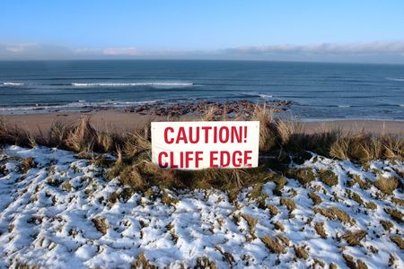 a red caution sign on a cliff edge in snow covered ballybunionの写真素材