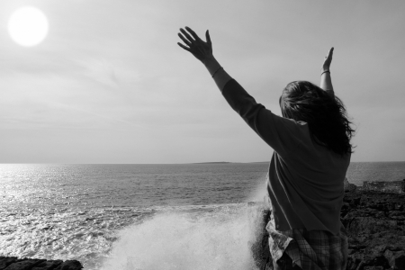 a lone woman raising her arms in awe at the powerful waves on the cliffs edge in county clare ireland in black and whiteの写真素材