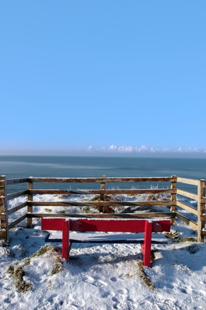 a seasonal snow frosty covered red bench with a sea view and blue cloudy sky background with copy spaceの写真素材