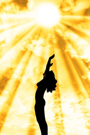 attractive silhouetted nude woman holding her hands up to the sky giving gratitude to the heavens in a yoga pose with a cloudy backgroundの写真素材